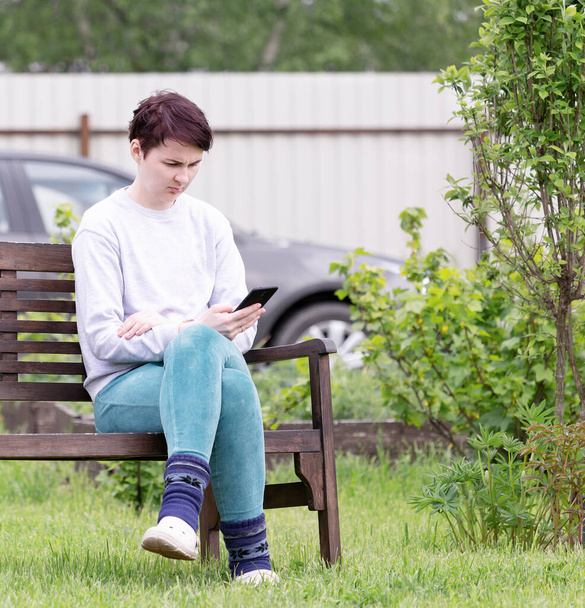 Ένα κορίτσι με ρούχα στο σπίτι κάθεται σε ένα παγκάκι με ένα smartphone στο χέρι της - Φωτογραφία, εικόνα