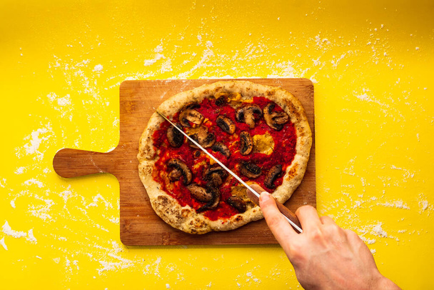 Männliche Hand schneidet vegane, rustikale hausgemachte Pilzpizza auf einem Holzschneidebrett auf gelbem Hintergrund - Foto, Bild