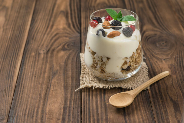 Yogur, granola y bayas en un vaso y una cuchara de madera en una mesa de madera
. - Foto, Imagen