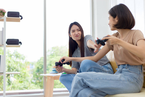 Twee jonge Aziatische vrouwen die wedstrijdjoysticks vasthouden en vrolijk op de bank zitten. Concurrerende vrienden spelen videospelletjes thuis. - Foto, afbeelding