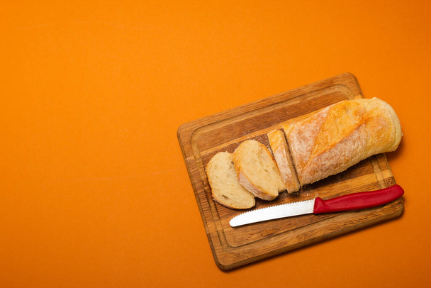 Κομμένη μπαγκέτα ψωμιού σε ξύλινο ξύλο κοπής με μαχαίρι σε φόντο χρώματος τερακότ. Αντιγραφή χώρου. Κοντινό πλάνο υψηλής ποιότητας φωτογραφία - Φωτογραφία, εικόνα