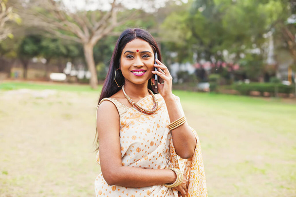 Beautiful indian woman in saree talking on mobile phone, standing in the park - 写真・画像