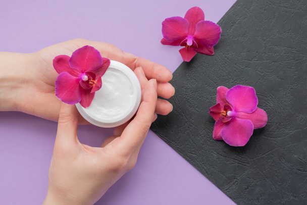 Cosmetic background with hands, soft white cream and fragile magenta colored orchids.Moisturizing facial cream in a jar and blooming orchid flowers on purple and black background, skin care cosmetics. - Foto, Bild