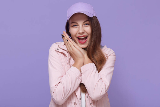 Portrait of glad full of happiness girl with wide open mouth and happy eyes, keeping palms together near cheek, looking at camera isolated on lilac background. - Photo, image