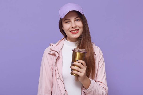 Shot of glad female student has coffee break after lectures, enjoys hot beverage from thermo mug, posing isolated over lilac background, wearing casual attire. - Foto, Imagen