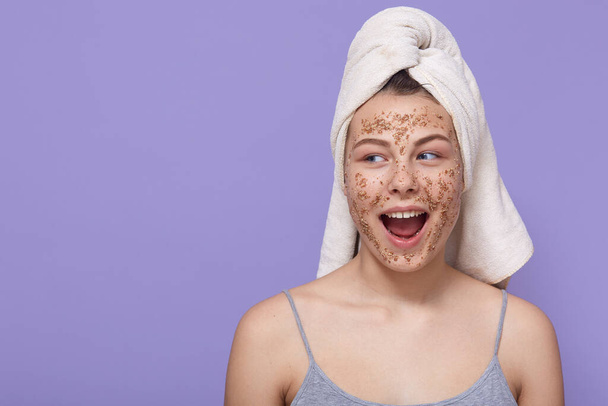 Surprised girl wearing white towel doing morning procedures, having facial mask, posing with widely opened mouth against lilac wall, female moisturize skin. - Фото, изображение