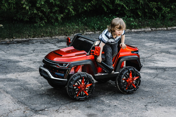 ストライプのTシャツを着た巻き毛の少年は、アスファルトの道を走る赤い大きなおもちゃの車に乗っています。屋外でのレクリエーションは - 写真・画像