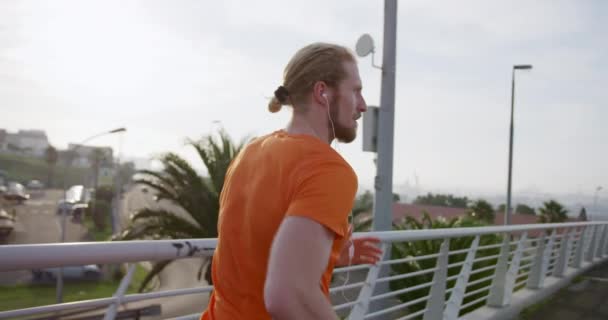 A young athletic Caucasian man exercising on a footbridge in a city, running and listening to music with earphones on in slow motion. Healthy urban active lifestyle. - Video, Çekim