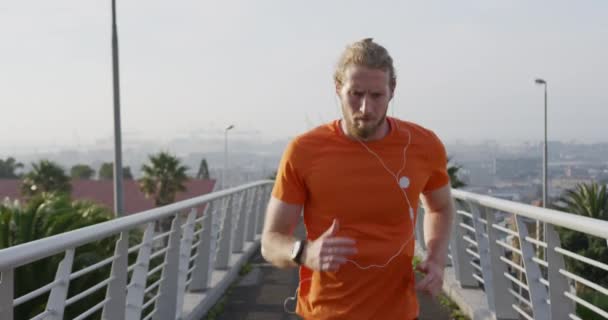 A young athletic Caucasian man exercising on a footbridge in a city, running and listening to music with earphones on in slow motion. Healthy urban active lifestyle. - Záběry, video