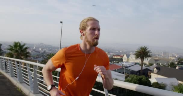 A young athletic Caucasian man exercising on a footbridge in a city, running and listening to music with earphones on in slow motion. Healthy urban active lifestyle. - Πλάνα, βίντεο