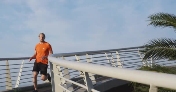 A young athletic Caucasian man exercising on a footbridge in a city, running and listening to music with earphones on in slow motion. Healthy urban active lifestyle. - Séquence, vidéo