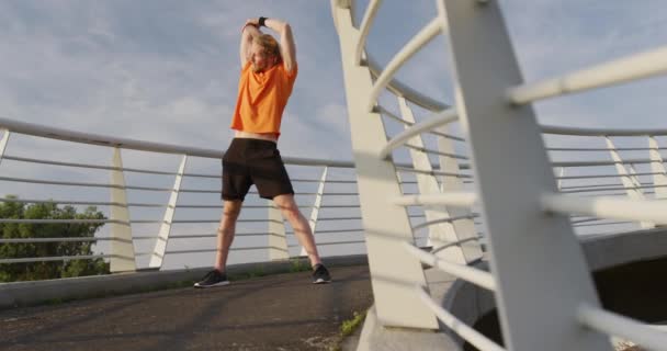 A young athletic Caucasian man exercising on a footbridge in a city, stretching out in slow motion. Healthy urban active lifestyle. - Кадри, відео