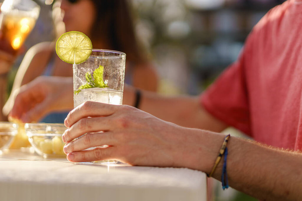 Mojito mit Zitronenscheibe an einem Restauranttisch. Aus nächster Nähe auf das Glas und eine männliche Hand. Menschen, die alkoholische oder alkoholabhängige Getränke trinken. - Foto, Bild