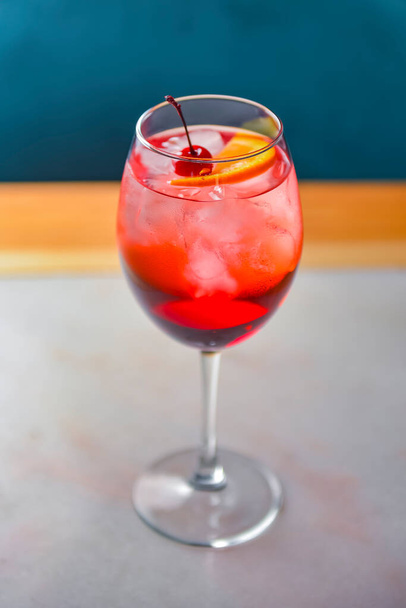 A glass with the red citrus cocktail. Alcohol summer beverage in a wine glass. Refreshing drink with ice on a wooden table inside a restaurant. Eating out concept. Blurred background. - Foto, Bild