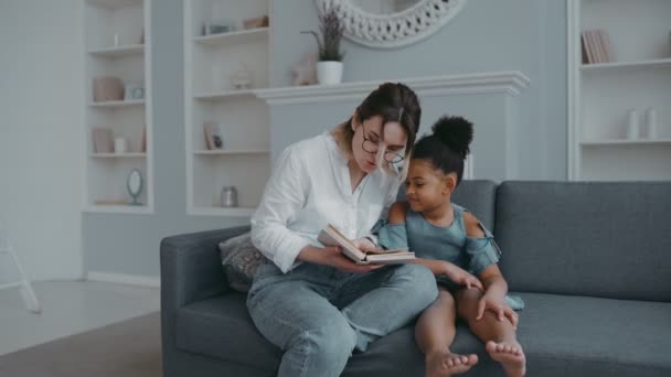 Young womans, mother, foster parent or teacher reading book to her little african daughter. Girl listens carefully, smiling, sitting on sofa in living room. Education, teaching, preschool education - Metraje, vídeo