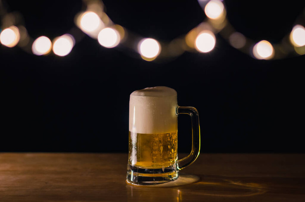 A glass of beer on wooden table that have bokeh lights on top with dark background. - Photo, Image