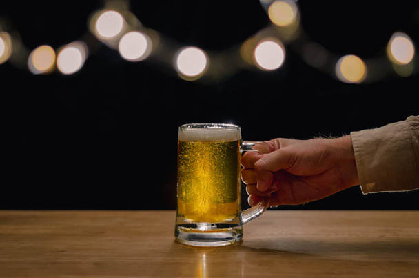 Mano sosteniendo un vaso de cerveza en la mesa de madera que tiene luces bokeh en la parte superior con fondo oscuro
. - Foto, imagen