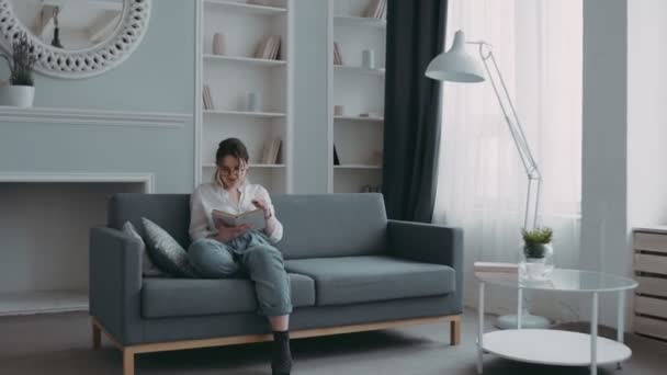In stylish, modern, bright living room, young european mother is reading book on sofa. Little beautiful african daughter runs up playing and gives her strong hug. Happy mother joyfully kisses girl - Filmmaterial, Video