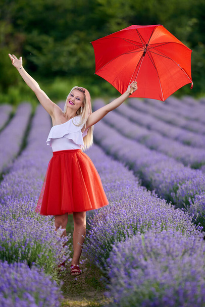 Beautiful young woman with red umbrella and skirt in a lavender plantation on rows - 写真・画像