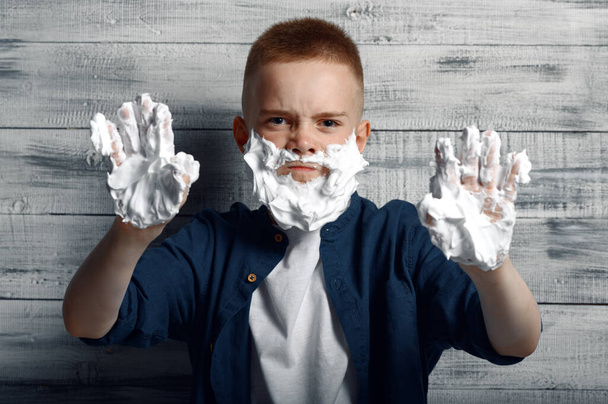 Little boy with shaving foam on his face and hands in studio. Kid isolated on wooden background, child emotion, schoolboy photo session - Фото, зображення