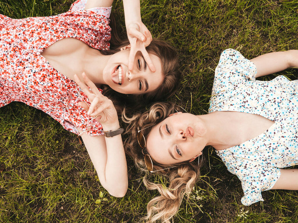 Two young beautiful smiling hipster girls in trendy summer sundress.Sexy carefree women lying on the green grass in sunglasses.Positive models having fun.Top view.They show peace sign - Φωτογραφία, εικόνα