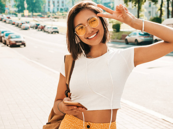 Fashion portrait of young stylish hipster woman walking in the street.Girl wearing cute trendy outfit.Smiling model enjoy her weekends, travel with backpack. Female listening to music via headphones - Photo, image
