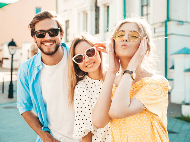 Group of young three stylish friends posing in the street. Fashion man and two cute girls dressed in casual summer clothes. Smiling models having fun in sunglasses.Cheerful women and guy at susnet - Zdjęcie, obraz