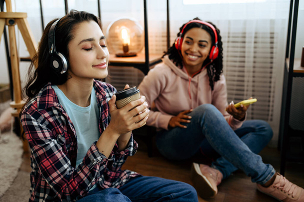 Two cute women enjoys listening to music with coffe at home. Pretty girlfriends in earphones relax in the room, sound lovers resting on couch, female friends leisures together - Photo, Image