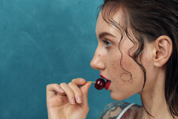 Nahaufnahme Porträt eines jungen Modells mit Kirschbeere. Sommersprossige Mädchen berührt Lippen mit Kirsche vor blauem Hintergrund. - Foto, Bild