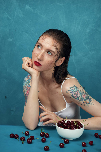 Verträumtes weibliches Modemodel sitzt am Tisch mit Kirschbeeren vor blauem Hintergrund  - Foto, Bild