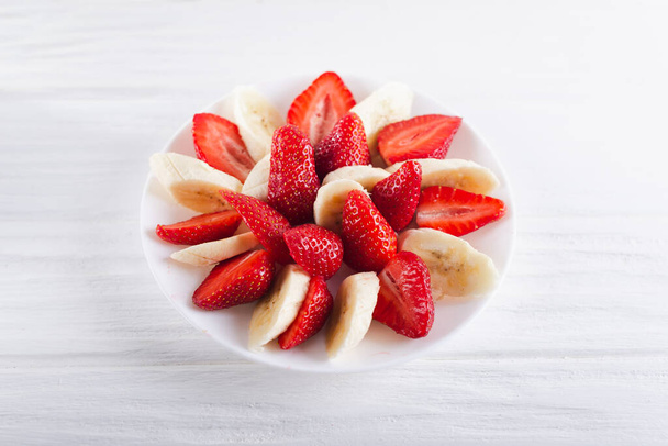 Juicy fresh sliced strawberries with banana on a white plate on a white wooden background - Fotó, kép