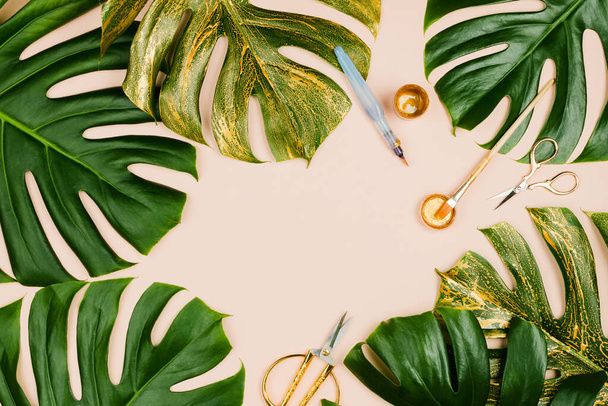 Monstera leaves painted golden with various stationary accessories: brushes, scissors, paint on pastel background - Φωτογραφία, εικόνα
