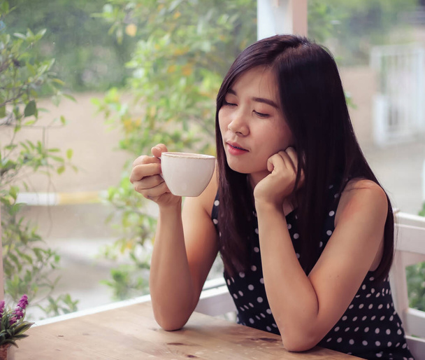 Kaunis aasialainen nainen istuu kahvilassa ikkunan vieressä pitäen kahvikuppia, sulkien silmänsä ja nauttien kahvin tuoksusta.. - Valokuva, kuva