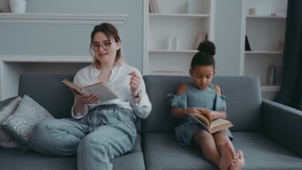 Portrait of modern young mother with an african foster daughter in living room. Woman teacher reads on sofa, and child listening with book in his hands. Concept of happy family or preschool education - Кадры, видео