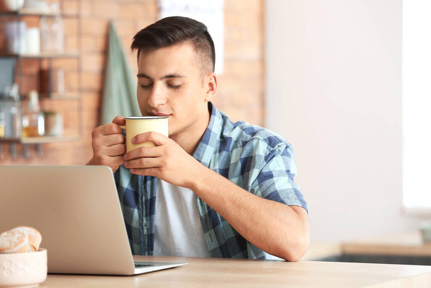 Komea nuori mies kannettavan tietokoneen juominen teetä kotona - Valokuva, kuva