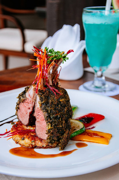 Pečený jehněčí steak s křupavou bylinkovou kůrkou a grilovanou zeleninou na bílém talíři s modrou margaritou v pozadí - Fotografie, Obrázek