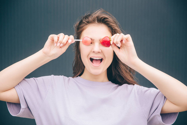 Young stylish trendy woman isolated over grey blue background. Funny positive girl posing on camera covering eyes with one lollipop and strawberry. Having fun. - Photo, Image