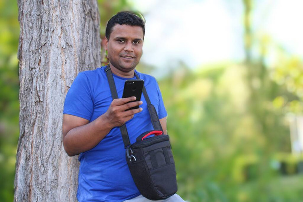 Mavi tişörtlü genç adam ağaçla parkta cep telefonu kullanıyor. - Fotoğraf, Görsel