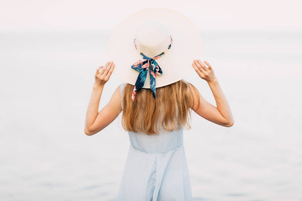 Back view, an attractive girl in a dress and a summer hat, resting and admiring the sea with her back to the camera, on the sea. Summer vacation, travel - Φωτογραφία, εικόνα