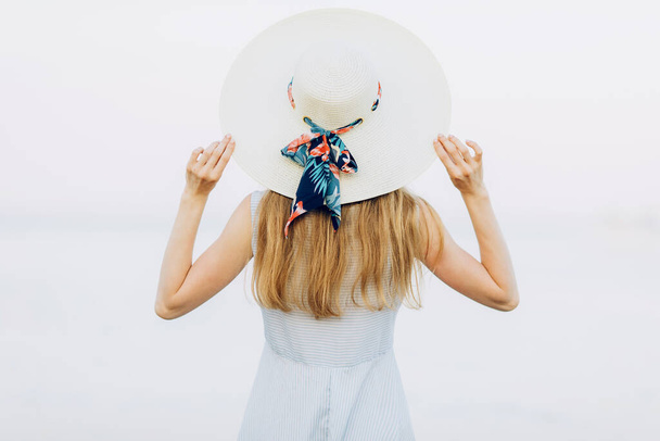 Back view, an attractive girl in a dress and a summer hat, resting and admiring the sea with her back to the camera, on the sea. Summer vacation, travel - Foto, immagini