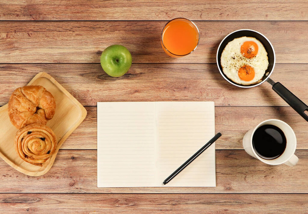 Widok góry zdrowy zestaw śniadaniowy, notatnik i długopis na drewnianym stole. Ciasto rogalikowe i duńskie, jabłko, sok pomarańczowy, smażone jajka na patelni, filiżanka czarnej kawy i notatnik. Morning rutynowe concept.planning swój dzień. - Zdjęcie, obraz