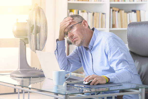 Ο άνθρωπος υποφέρει από θερμότητα ενώ εργάζεται στο γραφείο και προσπαθεί να δροσιστεί από τον ανεμιστήρα - Φωτογραφία, εικόνα