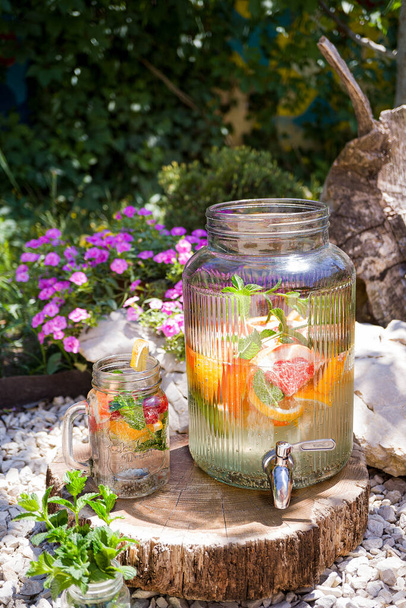 Refreshing lemonade with grapefruit, lemon, lime and mint in a glass jug with a tap and a mug is standing outdoors in the garden. The concept of drinks, summer, relaxation, healthy food. Vertically - 写真・画像