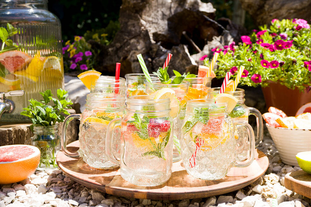 Infused water with lemon, grapefruit, lime and mint in glass cocktail mugs stands on a rotating wooden tray in the open air. Sunny day, bright pink and raspberry flowers in the background, relaxation. - 写真・画像