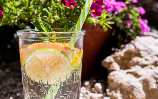 One glass glass with a cool drink, fruit slices and mint stands outdoors in the garden. In a glass is a striped green cocktail straw. Bright pink flowers in the background. Sunny day, vacation. Close-up - Фото, изображение