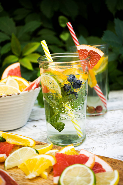 One glass with an ice drink with fruits and berries stands on a wooden table against a background of green foliage. Near a wooden cutting board with fruit slices (lemon, lime, orange, grapefruit). Vertically - Photo, Image