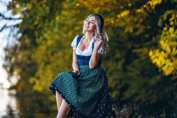 Oktoberfest, St. Patrick day, intenational beer day. Happy pretty blond girl in dirndl, traditional beer festival dress, sitting outdoors with blured colorful trees behind - Foto, imagen