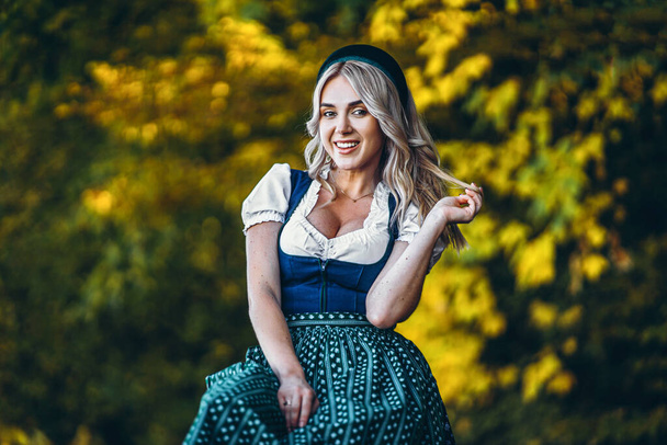 Oktoberfest, St. Patrick day, intenational beer day. Happy pretty blond girl in dirndl, traditional beer festival dress, sitting outdoors with blured colorful trees behind - Fotografie, Obrázek