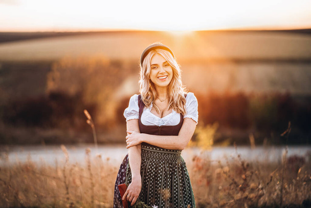 Pretty happy blonde in dirndl, traditional festival dress, sitting with two mugs of beer outdoors in the field with blurred background. Oktoberfest, St Patricks day, international beer day concept. - Foto, afbeelding