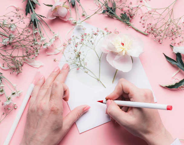 Female Hands write on envelope with flowers on pink tables background.  Wedding, invitation ,Valentine day,  Mother's Day greeting and love concept. Flat lay, top view, copyspace - Fotoğraf, Görsel